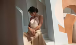 a woman in a tan outfit sits against an abstract structure