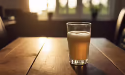 a glass of beer on a wooden table with the sun shining through the window