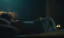 a woman lying in bed, dressed in blue, is illuminated by the lamp
