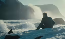 a man is sitting on snow near the ocean
