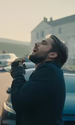 a bearded man outside of a house, standing with his hand over his mouth