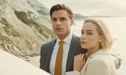a man in a suit and a woman standing close together by the ocean