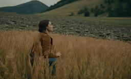 a woman stands in tall brown grass looking away