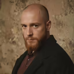 man with goatee standing against a wall and looking into camera