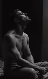 a shirtless man sits on the edge of his bed
