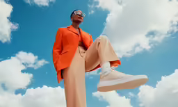a woman with a bright orange blazer and pants, is holding a sneakers up to her feet
