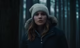 a woman in a parka standing in front of a forest