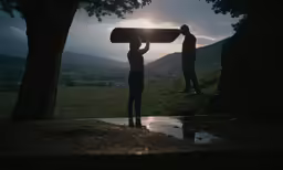 two men are standing under the shade of trees with a skateboard on their shoulders