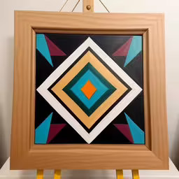 a brown frame with some bright shapes painted on it,colorful artwork with an open frame on a table,this is a painting on top of a white table,a painting on a wall with one block cut out of it and the other made up of it,the painting is on the easel above the table