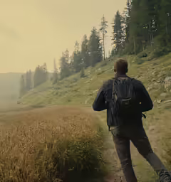 man walking through the grass in front of a hilly hill