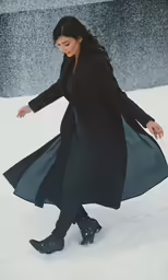 a woman in long coat, dress and snow boots walking across snow