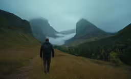a man hiking a grassy trail towards the mountains in fog