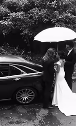 a bride and groom standing under an umbrella by a car