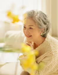 a woman smiling for the camera in front of a yellow flower,an older lady is wearing a white sweater and smiling at the camera