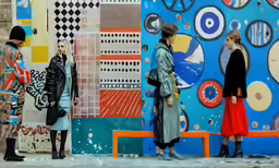 three young women stand on the sidewalk in front of a colorful wall with a mural
