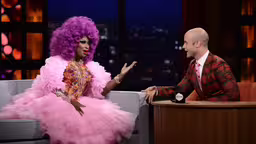 the man in the suit has pink hair and purple wigs on,a bald man in a suit and pink dress sitting next to an ope dressed up woman