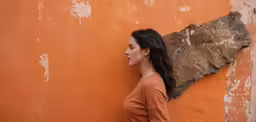 a girl with long hair leaning on an orange wall