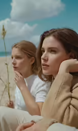 two women sit on the sand together, one holding her head