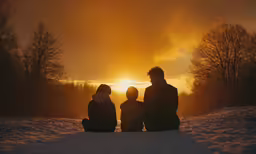 a group of three people sit together in the snow as the sun rises