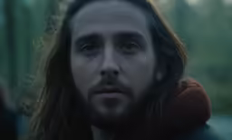 a man with long hair and blue eyes standing in a forest