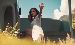 a woman in a white dress sitting down holding a hand out to a car