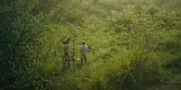 two people walking through the brush, in a large area with grass