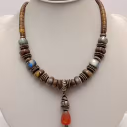 necklace that has various colored beads on it,a necklace has beads with wood beads on it,an elaborate necklace is made with beads and stones,a white mannequin with brown and orange beads,necklace with brown beads and colorful items on display
