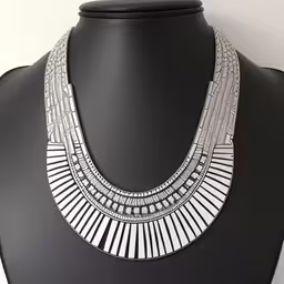 a white necklace is sitting on a black mannequin,a necklace that is set on top of a table,a necklace made with two layers of metal and glass,the egyptian statement necklace is worn in white and silver,a silver necklace is displayed on a mannequin
