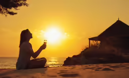 a woman sits on the beach with a wine glass and watches the sun go down