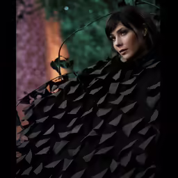 a model with an embellished cape stands near a bush