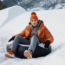 a woman wearing an orange coat sitting on a snow bag in the snow