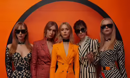 four women stand in front of a wall wearing blazers and high heels