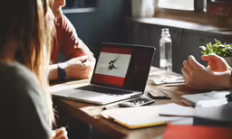 two women and a man in a red shirt sitting at a table with an open laptop