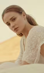 a woman in a white shirt looks at something while sitting on top of the sand dunes