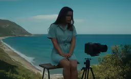 woman sitting on bench taking photo with camera next to ocean