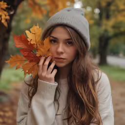 a young woman in winter clothing holds up an autumn leaf
