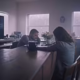 a man sits at a kitchen table with a cup on the table as a woman sits and watches