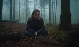 a young woman sitting in the middle of a forest in front of a tree stump