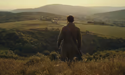 a man in a trench coat looking out over the hills