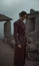 a woman in a brown dress and long trench coat with two stone buildings in the background
