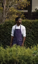 a man in an apron is standing outside near a hedge