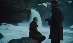 a man standing next to a woman sitting on top of a rock