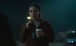 a woman with a cup of coffee standing in the middle of a dark street