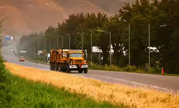 there are cars driving down the road near some trees,there is a yellow garbage truck parked along the side of the road