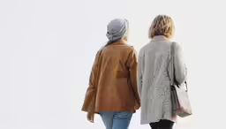 two women walking together, one is wearing a brown jacket and the other is a tan top