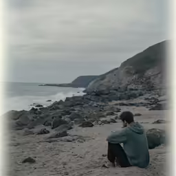 a man sits on the sand next to a body of water