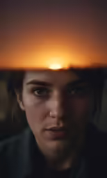 a woman stares into a mirror with the sun setting behind her