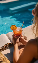 someone sitting at a table by a swimming pool holding a beverage in front of a book