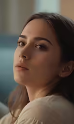a woman with brown hair and light blue eyes looks off to the side