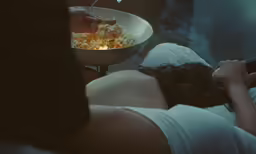 person laying on bed, eating popcorn and drinking wine